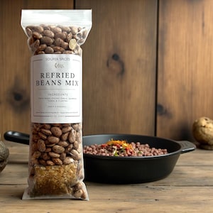 May include: A clear plastic bag of Souper Spices Refried Beans Mix. The bag contains pinto beans, spices, and the ingredients are listed on a white label. A black pan with refried beans is in the background. The bag is 8 oz (226g).