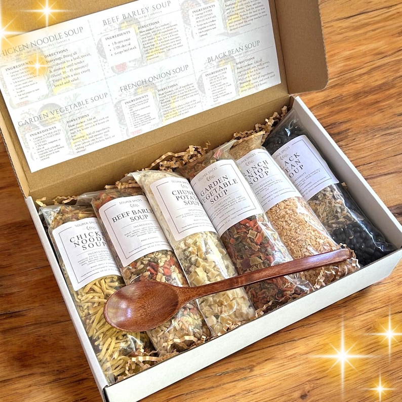 May include: A cardboard gift box containing six clear plastic pouches of dried soup mixes, including Chicken Noodle, Beef Barley, Chunky Potato, Garden Vegetable, French Onion, and Black Bean. A dark wooden spoon rests across the pouches.