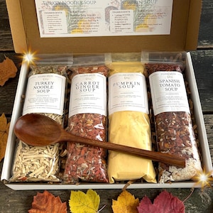 May include: A cardboard gift box containing four clear plastic pouches of dry soup mixes labeled Turkey Noodle, Carrot Ginger, Pumpkin, and Spanish Tomato Soup, with a long wooden spoon resting horizontally across the front of the pouches.