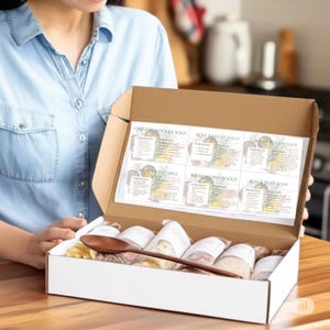 May include: A white cardboard box containing several small, labelled ingredient packets for various soups and a dark wooden spoon. The inside lid features printed recipe cards for Chicken Noodle, Beef Barley, Chunky Potato, Garden Vegetable, French Onion, and Black Bean soups.