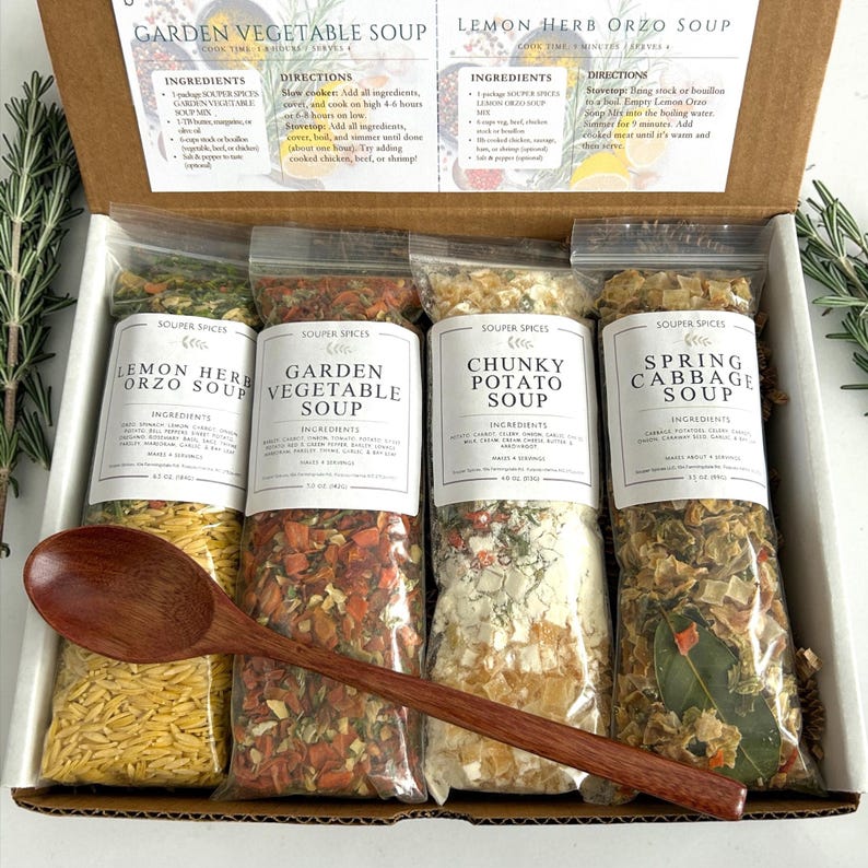 May include: A box set of four soup mixes: Lemon Herb Orzo, Garden Vegetable, Chunky Potato, and Spring Cabbage. Each packet is labeled with ingredients. A wooden spoon rests across the packets. The box includes instructions for slow cooker and stovetop methods.