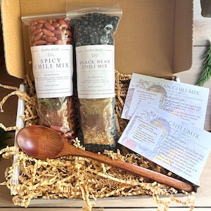 Könnte beinhalten: Eine Geschenkbox mit zwei Beuteln Chilimix, einem scharfen und einem mit schwarzen Bohnen, mit Zutaten- und Anweisungskarten. Ein Holzlöffel liegt auf dem geschredderten Papier. Die Beutel sind mit "Souper Spices" beschriftet.