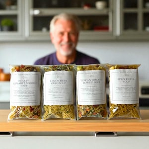 May include: Four clear plastic bags of soup mixes are displayed, each with a white label. The labels read "Hearty Alphabet Noodle Soup", "Lemon Herb Orzo Soup", "Tuscan Tomato Bowtie Soup", and "Spicy Fiesta Soup". A person is blurred in the background.
