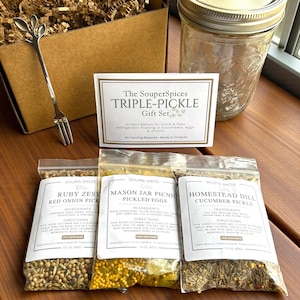Op de afbeelding: Een cadeaupakket met een decoratieve vork, een glazen pot en een doos met een kaart met de tekst "The SouperSpices TRIPLE-PICKLE Gift Set". De set bevat drie kruidenpakketten voor het inmaken van komkommers en eieren.