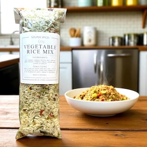 May include: A clear bag of Souper Spices Vegetable Rice Mix next to a white bowl of cooked rice. The bag has a label with the product name and ingredients. The bowl of rice is steaming and contains vegetables.