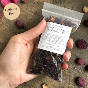Puede incluir: Una bolsa de plástico transparente llena de té de hierbas seco, sostenida en una mano. La etiqueta dice "Berry Citrus Herbal Tea" con una lista de ingredientes. Una pegatina redonda indica "Sin cafeína". El té parece ser una mezcla de frutas y hierbas secas rojas y marrones.