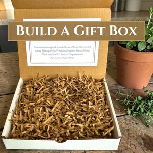 May include: A brown cardboard gift box filled with shredded brown paper filler. The inside lid features a white card with the text BUILD A GIFT BOX and a list of custom message options including Thinking of You, Sympathy, and Birthday.
