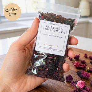 May include: A clear plastic resealable pouch filled with dried dark red hibiscus tea leaves. The white label reads: SOUPER SPICES RUBY-RED HIBISCUS TEA, ORGANIC HIBISCUS, CAFFEINE FREE, 10 SERVINGS - 1 TB, 16 OZ (90G). A round beige sticker says Caffeine Free.