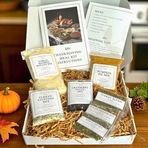 May include: A Thanksgiving meal kit in a white box, containing spice mixes, instructions, and a menu. The kit includes mixes for garlic mashed potatoes, turkey stuffing, cranberry sauce, and pumpkin pie. A small pumpkin and fall leaves are nearby.