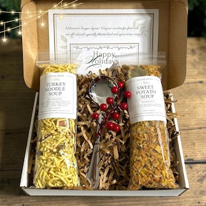 May include: A cardboard gift box containing two clear plastic bags of dried soup mix labeled Turkey Noodle Soup and Sweet Potato Soup, a silver metal soup spoon, and a decorative sprig of red berries, all nestled in brown paper filler.