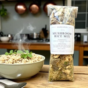 May include: A bowl of cooked mushroom rice with fresh herbs sits next to a package of mushroom rice mix. The package label reads "Mushroom Rice Mix" with ingredients and directions. The background shows a kitchen setting.