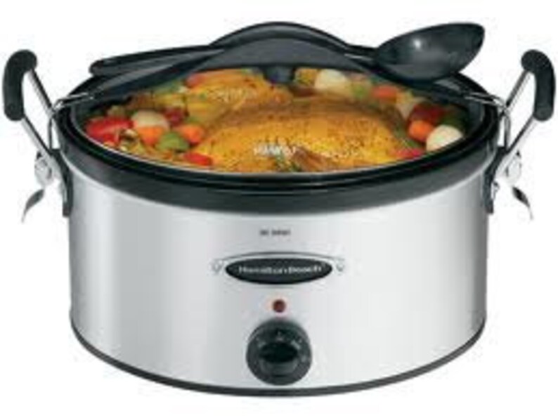 May include: A silver slow cooker with a black lid and a black handle. The slow cooker has a round shape and a black dial on the front. The slow cooker is filled with food, including a whole chicken, vegetables, and onions.