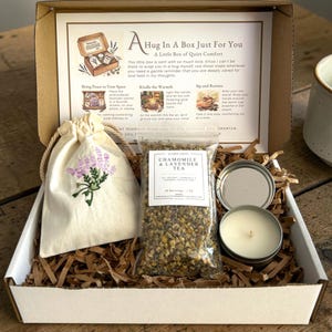 May include: A gift box containing a cream-colored drawstring pouch with lavender embroidery, a clear bag of chamomile and lavender tea, and a small round candle in a silver tin. An instructional card titled A Hug In A Box Just For You is included.