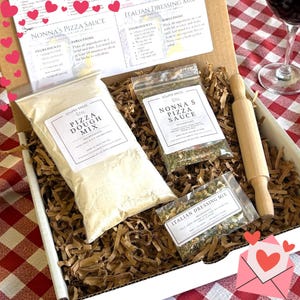 May include: A cardboard gift box containing a bag of pizza dough mix, a bag of Nonna's pizza sauce mix, a bag of Italian dressing mix, and a small wooden rolling pin. The box includes printed recipe cards for pizza sauce and dressing.