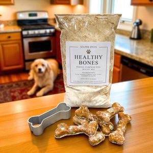 May include: A clear plastic bag of Souper Spices Healthy Bones Pawkin Pumpkin dog biscuit mix, 9.0 oz, with a metal bone-shaped cookie cutter and several baked bone-shaped dog treats on a wooden surface. The label lists ingredients and yield.