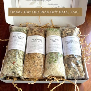 May include: A gift box containing four clear plastic bags of rice mixes: Vegetable, Spanish, Risi Bisi, and Mushroom. Each bag has a white label with the product name and ingredients. The box includes gold-colored paper filler and the text "Check Out Our Rice Gift Sets, Too!"