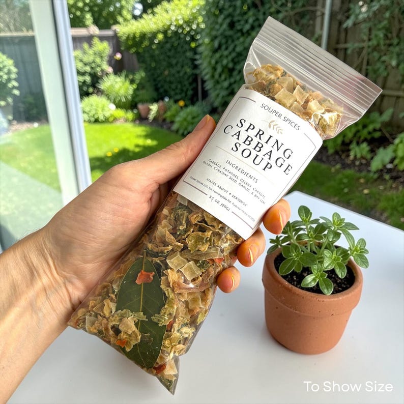 May include: A clear plastic bag of Souper Spices Spring Cabbage Soup mix. The label lists ingredients and says it makes about 4 servings. A small terracotta pot with a green plant sits nearby. The bag contains 3.5 oz of soup mix.