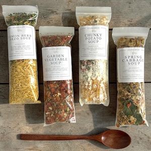 May include: Four clear plastic bags of soup mixes labeled "Lemon Herb Orzo Soup", "Garden Vegetable Soup", "Chunky Potato Soup", and "Spring Cabbage Soup". A wooden spoon rests below the bags on a wooden surface.