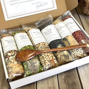 May include: A gift box filled with various soup mixes, including Heart Bean, Split Pea, and Black Bean. Each mix is packaged in a clear bag with a white label. A wooden spoon rests across the bags. The box also contains a handwritten note.