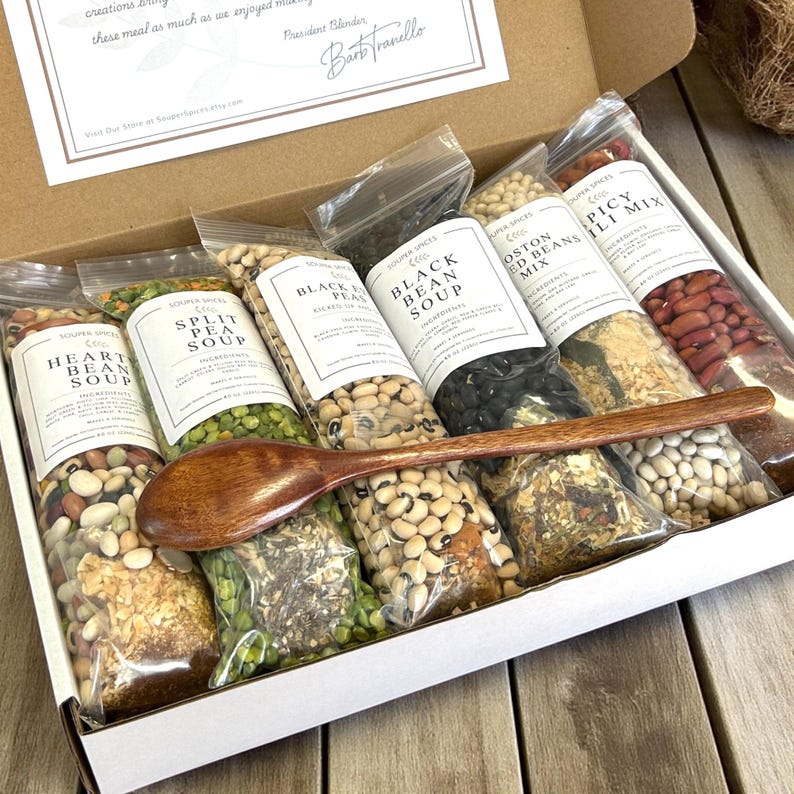 May include: A gift box filled with various soup mixes, including Heart Bean, Split Pea, and Black Bean. Each mix is packaged in a clear bag with a white label. A wooden spoon rests across the bags. The box also contains a handwritten note.