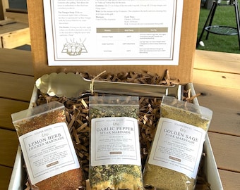 May include: A cardboard gift box containing three clear plastic pouches of dry spice blends labeled Lemon-Herb Chicken Marinade, Garlic Pepper Steak Marinade, and Golden Sage Pork Marinade, along with silver metal tongs and an instructional card.