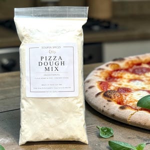 Puede incluir: Una bolsa de plástico transparente de mezcla para masa de pizza con el texto "PIZZA DOUGH MIX" en la etiqueta. La bolsa está al lado de una pizza cocinada con pepperoni y albahaca.
