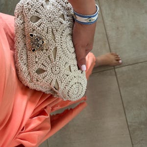 May include: A white crocheted bag with a gold animal charm. The bag is being held by a person wearing a coral colored dress and a blue and white bracelet.