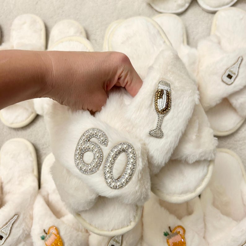 May include: White faux fur slippers featuring a beaded number 60 patch and a beaded champagne flute patch. The slippers have a soft, plush texture and a cross-band design. The number 60 is outlined in small white pearls, while the champagne flute is detailed with gold and silver beads.