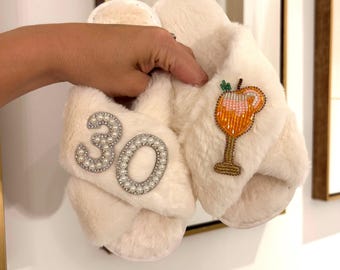 May include: A pair of cream-colored faux fur slippers with crisscross straps. One strap features the number 30 outlined in white pearls and clear crystals. The other strap displays a beaded cocktail glass patch in orange, gold, and green.