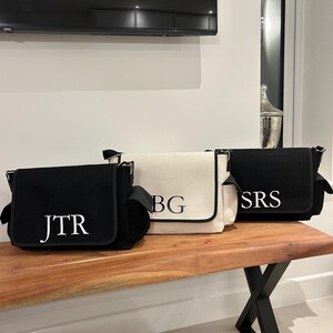 May include: Three canvas messenger bags with black straps and white embroidered initials. The bags are lined up on a wooden surface. The initials are "JTR", "BG", and "SRS".