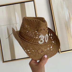 Könnte beinhalten: Brauner Cowboyhut aus geflochtenem Stroh mit einer perlenbesetzten "30" auf der Vorderseite. Der Hut hat eine breite Krempe und eine gewebte Textur. Ein Party-Accessoire.