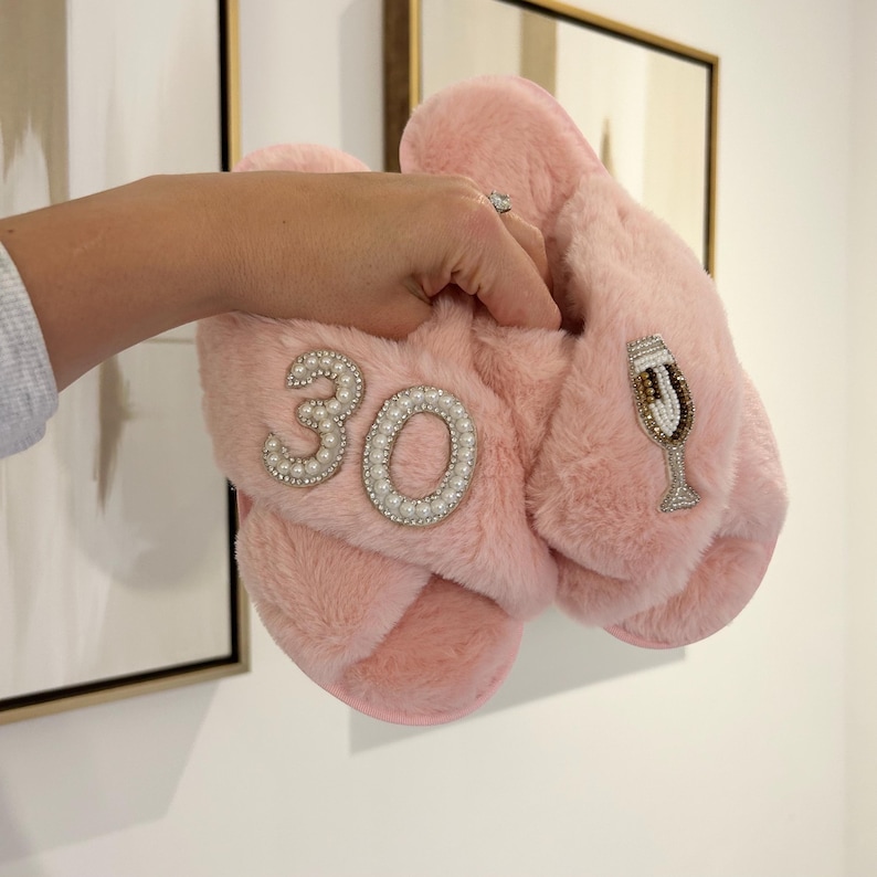 May include: A pair of light pink faux fur slippers featuring a cross-strap design. One slipper is decorated with the number 30 outlined in white pearls, while the other slipper features a beaded champagne flute appliqu&eacute; with gold and white accents.