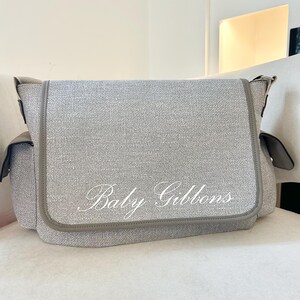 May include: A light gray baby bag with the text "Baby Gibbons" in white script. The bag has a flap closure and side pockets. The trim is a darker gray. The bag is sitting on a white surface.