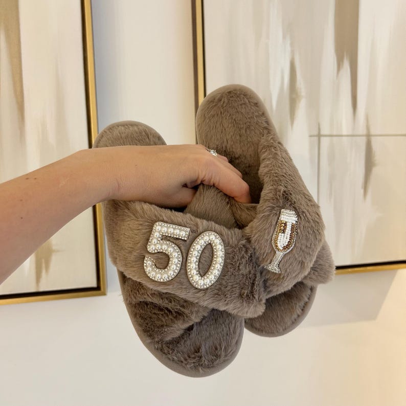 May include: A pair of taupe faux fur slippers featuring a crisscross strap design. One slipper is embellished with the number 50 outlined in white pearls, while the other features a decorative champagne flute patch accented with gold and white beads.