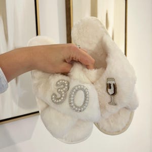May include: A pair of white faux fur slippers featuring a beaded design on the top straps. One strap displays the number 30 outlined in white pearls and clear crystals, while the other strap features a beaded champagne flute illustration.
