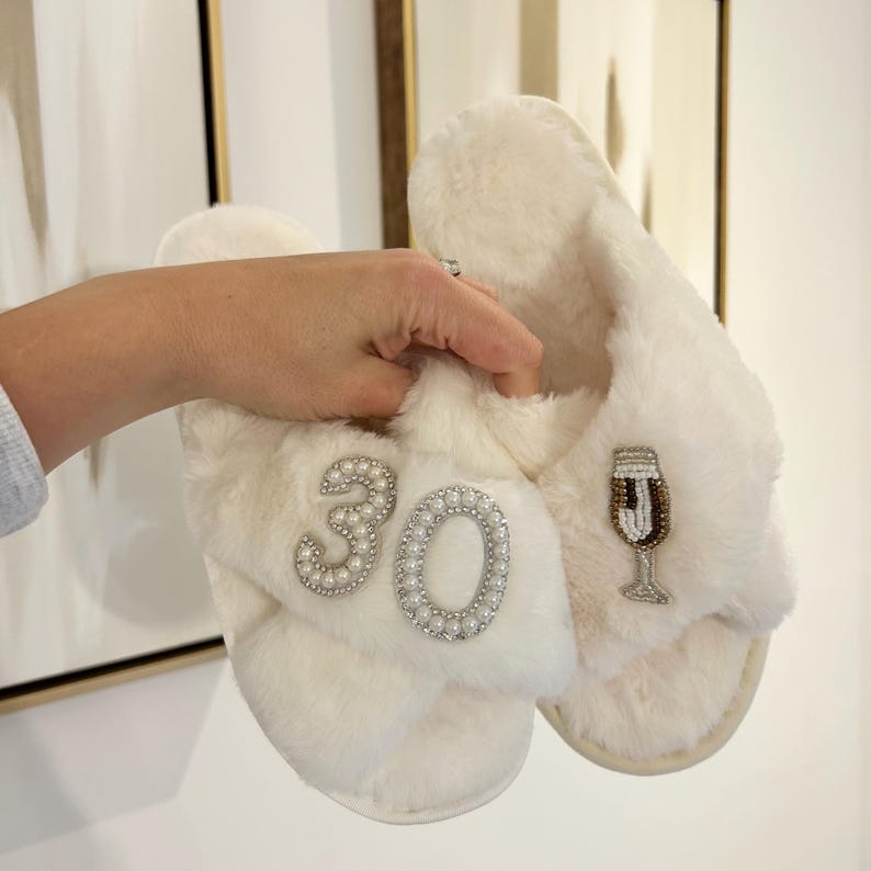 May include: A pair of white faux fur slippers featuring a beaded design on the top straps. One strap displays the number 30 outlined in white pearls and clear crystals, while the other strap features a beaded champagne flute illustration.