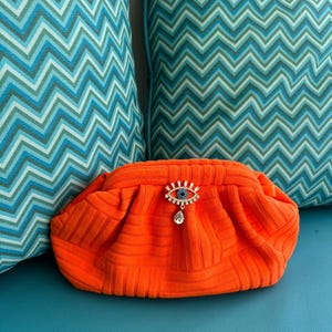 May include: Orange quilted clutch with a large, jeweled eye design. The eye is blue with white and clear rhinestones.