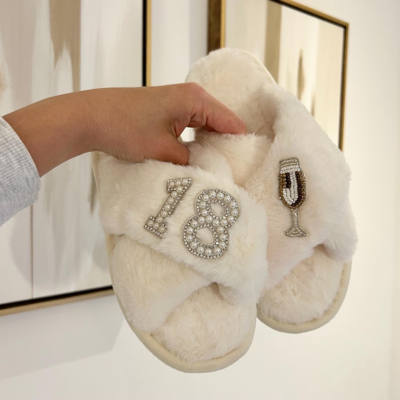 May include: A pair of cream-colored faux fur slippers featuring a crisscross strap design. One strap is embellished with the number 18 in white pearls and clear crystals, while the other strap displays a beaded champagne flute applique.