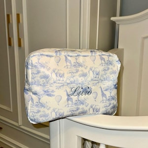 Op de afbeelding: Een wit en blauw toilettasje met ritssluiting. De tas heeft een safari-dierenprint met olifanten, giraffen en luchtballonnen. De naam "Livio" staat op de voorkant.