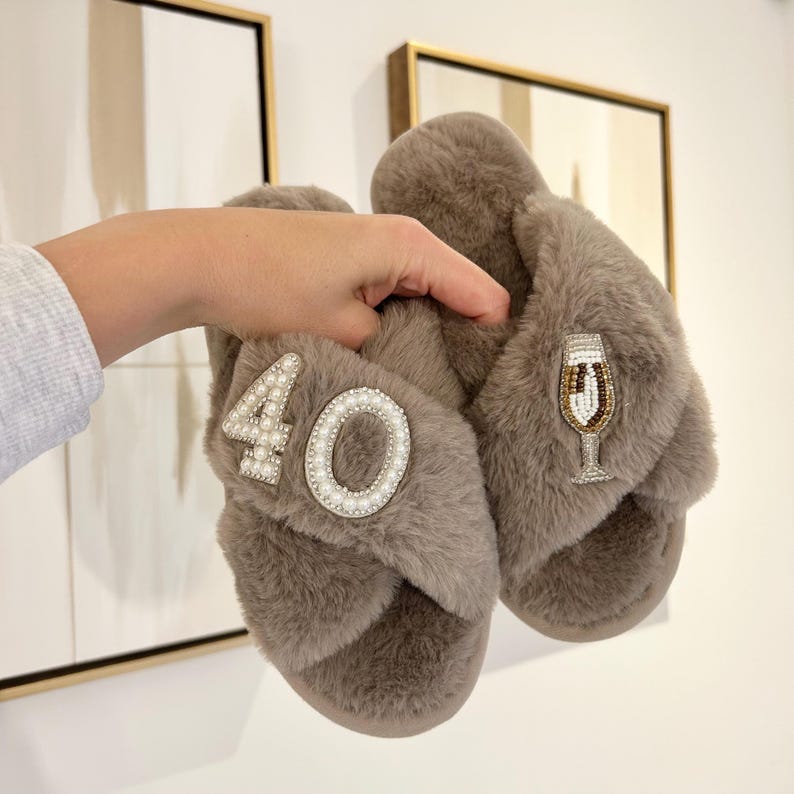 May include: A pair of taupe faux fur slippers with crisscross straps. One slipper features the number 40 embellished with white pearls, while the other displays a beaded champagne flute design with gold, white, and brown accents on the strap.