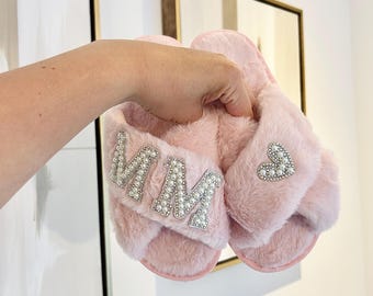 Può includere: Un paio di pantofole in pelliccia sintetica rosa chiaro con design a fasce incrociate. Una pantofola è decorata con le lettere MM in perle bianche e bordo argentato, mentre l'altra presenta una toppa a forma di cuore in perle bianche.