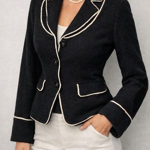 May include: A black blazer with cream-colored trim around the lapels, pockets, and cuffs. The blazer has a tailored fit, three buttons, and a classic design. The model is wearing a pearl necklace and cream-colored pants.