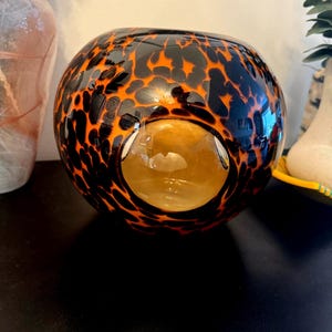 May include: A round glass bowl with a unique pattern. The bowl features a black and orange spotted design on the exterior, with a clear, golden interior. The bowl has a large opening on one side.