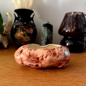 May include: A small, oval-shaped ceramic planter with a speckled brown and cream finish. The planter has two openings and sits on a wooden surface. Decorative vases and objects are in the background.