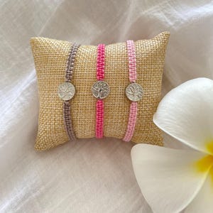 Può includere: Tre braccialetti in macramè con ciondoli argentati a forma di albero della vita sono esposti su un piccolo cuscino di iuta. I braccialetti sono nelle tonalità del marrone, del rosa e del rosa chiaro. Un fiore bianco è sullo sfondo.
