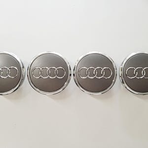 Könnte beinhalten: Vier graue Radnabenabdeckungen mit dem silbernen Audi-Logo. Jede Kappe hat eine silberne Umrandung und das silberne Logo mit den ineinandergreifenden Ringen. Die Kappen sind in einer Reihe vor weißem Hintergrund angeordnet.