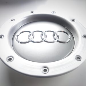 May include: Silver-colored car wheel center cap with the Audi logo. The cap is circular with a brushed metal finish and multiple bolt holes around the edge. The Audi logo consists of four interlocking rings.