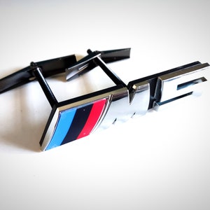 Könnte beinhalten: Ein silbernes BMW M Emblem mit einem blauen, schwarzen und roten Streifen. Das Emblem ist an einer schwarzen Metallhalterung befestigt.