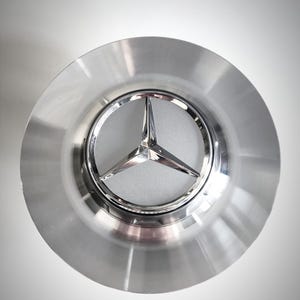 Op de afbeelding: Zilverkleurige wieldop met een verchroomd Mercedes-Benz logo in het midden. Het logo heeft een driepuntige ster in een cirkelvormig embleem. De wieldop heeft een geborstelde metalen afwerking en een ronde vorm.