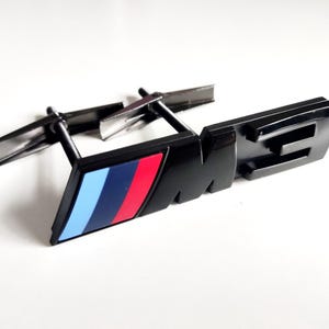 May include: Black plastic car emblem with a blue, red, and white stripe and the letters "M3" in raised lettering.